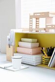 Architect's model above cardboard boxes and pen holder in yellow shelf unit with notebook and beaker on table in front