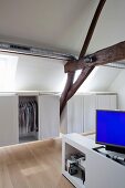 Clothes racks in custom floating wardrobes and blue monitor on foot of white bed in attic bedroom