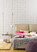 A double bed with a headboard and pillows next to a rustic swing used as a bedside table in front of grey patterned wallpaper