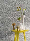 A yellow metal table with vases in front of non-woven wallpaper with a floral pattern in shades of grey