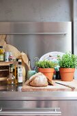 Bread on wooden chopping board and herbs in terracotta pots on stainless steel worksurface