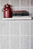 Red milk can and books on floating shelf on wall papered with book pages