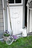 Lettering and sun made from bent wire on wooden door flanked by potted violas, spade and watering can