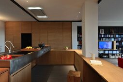 Open-plan, wooden designer kitchen with metal sink and integrated pillar; view of shelving in living area