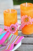 Pinkfarbener Streifenserviette und zwei orangefarbenen Kerzen mit Blüten und Geschenkbändern dekoriert