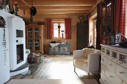Masonry wood-burning stove, elegant armchair, chest of drawers and farmhouse trunk in open-plan interior of wooden cabin