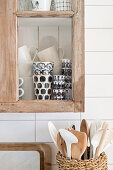 Basket of wooden spoons below black and white mugs in glass-fronted cabinet