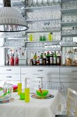 Küchenzeile mit verspiegelter Rückwand und vielen Gläsern auf Wandregal hinter gedecktem Esstisch mit bunten Gedecken