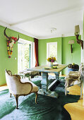 Solid wooden table, vintage benches covered with reindeer furs and velvet armchairs in green dining room with buffalo skull on wall