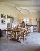 Long wooden dining table and chairs on concrete floor below fan on white, wood-beamed ceiling