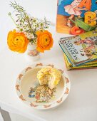 Posy in beaker, muffin with bite missing on child's plate in front of stacked of children's books