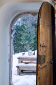 Blick durch rustikale, geöffnete Holztür auf Tisch und Bank im Schnee