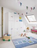 A white wardrobe with colourful handles and a hopscotch rug in a light and bright young girl's bedroom with slanted roof