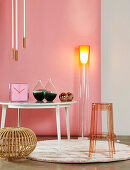 Designer standard lamp, clock and glass vases on table, transparent bar stool and rattan stool in front of pink wall