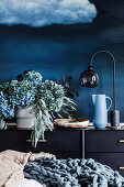 Vase mit Hortensien und Eucalyptuszweigen, Schale, Krug un Tischlampe auf Sideboard vor blauer Wand