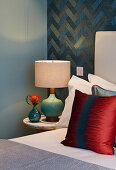 Bedside lamp next to bed with red scatter cushion against blue patterned wallpaper