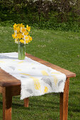 Vase of narcissus on table runner printed with pattern of feathers in garden