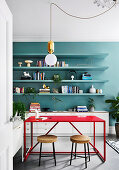 Red table with two stools and shelves on a petrol-colored wall in the study