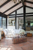 White blanket and cushions on rattan sofa and old trunk used as coffee table in conservatory with terracotta floor tiles