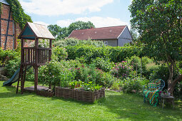 Holzhäuschen mit Kinderrutsche, Hochbeet aus Weidenästen und Sitzplatz unter Baum im Garten