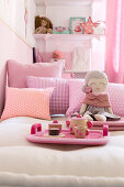 Tablett mit Puppengeschirr und eine Puppe auf dem Bett mit rosa Kissen