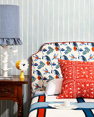 Bed with patterned headboard in front of stripe wallpaper, antique bedside table with table lamp and clock