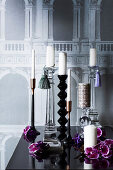 Various candle holders in front of a wallpaper with an architectural motif