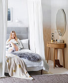 Woman reading in a comfortable canopy bed, a dressing table next door