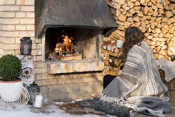 Woman sitting in front of open fire and stacked firewood outside