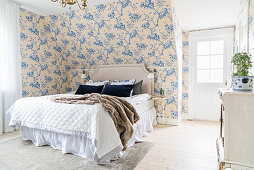 Double bed wiith headboard and chest of drawers in romantic bedroom with floral wallpaper