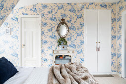 View across double bed to console table between door and fitted wardrobe in romantic bedroom with floral wallpaper
