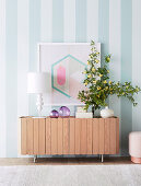 Sideboard with table lamp, glass decoration and flower sprig against striped wall