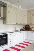 Simple, beige and white country-house kitchen with red-and-white striped runner adding a splash of colour