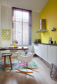 Classic rocking chair, table and white base units in kitchen with yellow wall and louvre blind on window