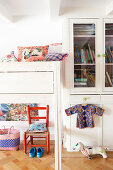 Colourful cushions on white loft bed next to glass-fronted cupboard in child's bedroom