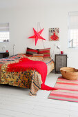 Colourful bedspread and red fringed blanket on double bed below red, star-shaped lamp on wall