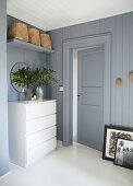 A vase of leafy twigs on a white chest of drawers next to a door in a blue-grey bedroom