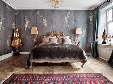 A double bed with an antique bed head flanked by floor lamps and an gold coloured statue in a bedroom