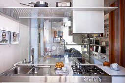 Blick über Edelstahl-Kücheninsel durch Glaswände in Wohnbereich mit Sofa und Schrankwand
