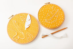 Instructions embroidering feathers on yellow fabric in embroidery frames