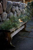 Wintry arrangement of conifer branches and candle lantern on wooden bench against stone wall