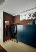 Clothes rail between tall, blue sideboard and mirror in bedroom with brown walls