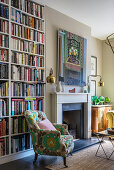 A floor-to-ceiling bookcase, an upholstered armchair and a modern work of art over the fireplace