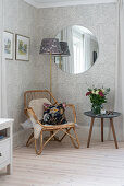 A rattan armchair, a side table, a floor lamp and a round mirror on a wall with patterned wallpaper