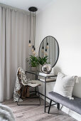 A dressing table, a round wall mirror, a pendant lamp and an upholstered bench in a bedroom