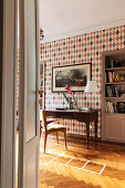 An antique desk against a wall with diamond-patterned wallpaper and a bookshelf in a wall niche