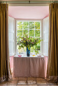 Tisch mit Blumenstrauß vor Fenster mit geöffneten Fensterläden, Vorhänge aus goldener Seid