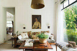 Sofa with cushions, portrait of woman above, table and bench in room with garden view