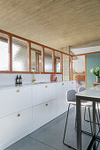 White, custom-made kitchen counter below glass wall and high table with bar stools