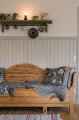 Wooden sofa bench with animal fur and cushions in front of beadboard paneled wall, above shelf with vintage decorative objects